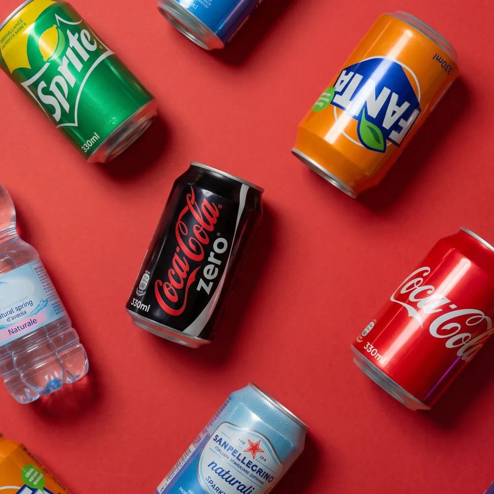 Lattine di bibite e una bottiglia d'acqua sparse su un fondo rosso, viste dall'alto