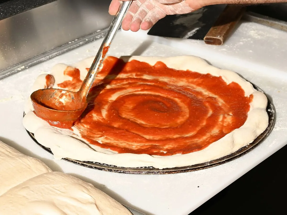La mano del pizzaiolo stende il pomodoro sulla pizza con il mestolo, accanto ai panetti che lievitano